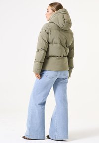 Chaqueta acolchada verde olivo con capucha, que presenta secciones acolchadas, bolsillos con cremallera y un diseño corto, combinada con jeans acampanados de color azul claro.