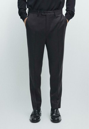 Mann trägt dunkle maßgeschneiderte Hose, schwarze Lederschuhe und ein schwarzes langärmeliges Hemd mit Händen in den Taschen vor neutralem Hintergrund.