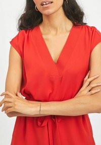 Blouse rouge à manches courtes avec décolleté en V, en tissu léger, présentant un détail noué à la taille et une texture lisse.