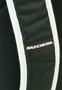 Tela negra texturizada con acentos blancos presenta un logo de "SKECHERS" en un parche liso. El diseño es deportivo y aerodinámico.