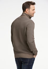 Brun stickad tröja med hög krage, mönstrad struktur och ribbad fåll, kombinerad med mörkblå jeans. Visas bakifrån.