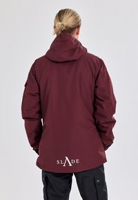 Maroon waterproof jacket with a hood, elastic cuffs, and front pocket. Features “SLADE” logo in white at the lower back. Textured fabric.