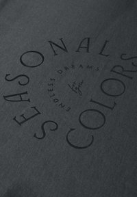 Dunkelgrauer Stoff mit fettgedrucktem schwarzem Schriftzug. Der Text umfasst "SAISONAL" und "FARBEN" und verfügt über eine glatte Textur sowie ein dezentes Muster.