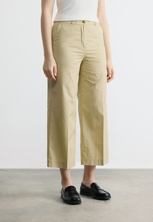 Άτομο που φοράει μπεζ φαρδιά παντελόνια μέχρι τον αστράγαλο και μαύρα μοκασίνια, όρθιο μπροστά σε απλό ανοιχτόχρωμο φόντο.