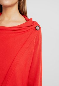 Femme portant un haut en maille rouge vif avec un col drapé maintenu par un grand bouton foncé sur l’épaule et des boucles d’oreilles créoles dorées visibles.