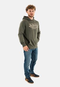 Sweatshirt vert olive avec poche kangourou à l'avant et logo, associé à un jeans en denim bleu et des baskets noires avec des accents beige.