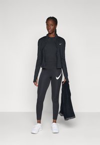 Kvinna i svart Nike träningsoverall och vita sneakers, hållande en svart handduk, stående mot en ljusgrå bakgrund.