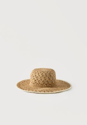Wide-brimmed, light brown crochet straw hat with a textured floral pattern and white trim along the edge, against a plain background.