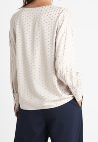 Blusa a maniche lunghe in beige chiaro con piccole pois dorati, caratterizzata da una vestibilità morbida, scollatura rotonda e polsi gathered.