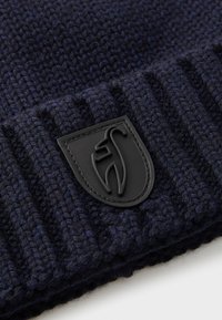Pletená čiapka v tmavomodrej farbe s textúrovaným rebrovaným vzorom. Na prednej strane sa nachádza čierna gumová nášivka s logom. Mäkkučký a teplý materiál.