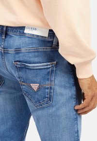 Jeans en denim bleu pour hommes avec un lavage clair, des coutures visibles et un patch en triangle emblématique de la marque GUESS sur la poche arrière.