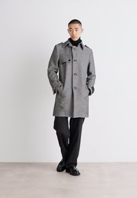Grå ullblandning kappa med krage och knappar, med en avslappnad passform och två sidofickor. Bärs över en svart polotröja och byxor.