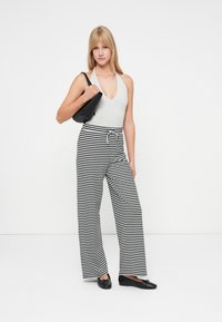 Body cinza com alças, calças largas às riscas pretas e brancas com cordão na cintura e sapatos pretos planos. O modelo segura uma bolsa preta.