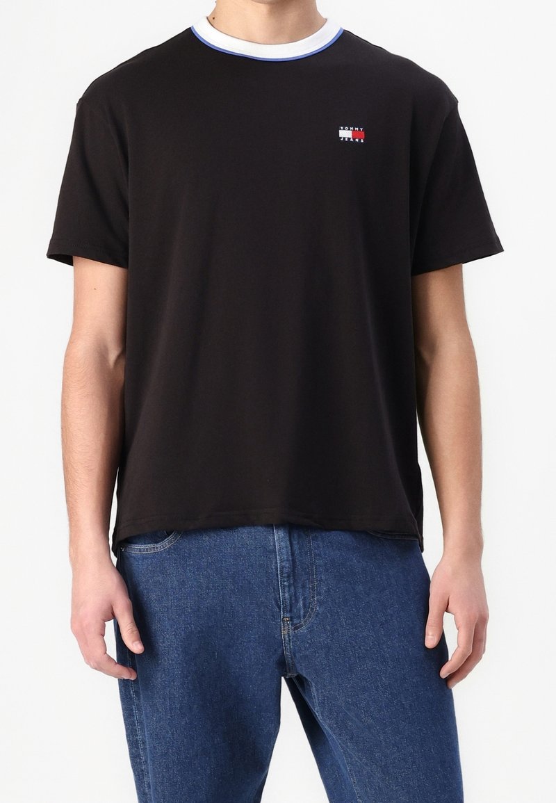 Mies, joka käyttää mustaa Tommy Jeans T-paitaa, jossa on sinireunainen valkoinen kaulus ja sinisiä farkkuja, näkyy kaulasta lantiolle valkoista taustaa vasten.