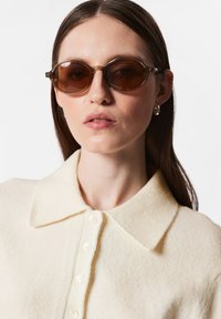 Braune ovale Sonnenbrille mit klaren Rahmen, kombiniert mit einem cremefarbenen, strukturierten Pullover mit einem Knopfkragen und vorderen Knöpfen.