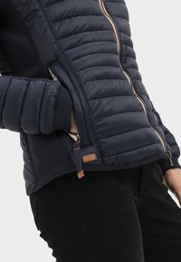 Navy puffer jacket with quilted sections, brown zipper, and leather tag. Features stretchy knit cuffs and a fitted design.
