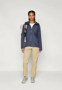 Casaco impermeável azul-marinho com fecho à frente, capuz ajustável e dois bolsos laterais, combinado com calças bege e ténis cinzentos.