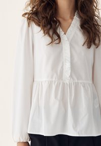 Woman wearing a white long-sleeve blouse with ruffled V-neckline and gathered waist, paired with black pants.