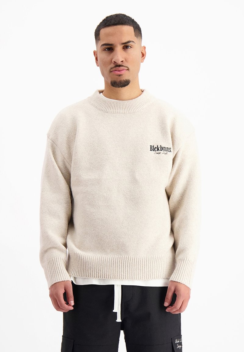 Crèmekleurige trui van zachte, gestructureerde stof, met een ronde halslijn en een zwart logo op de linkerkant van de borst. Geribbelde mouwen.