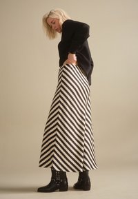 Blondynka poprawiająca czarny sweter, ubrana w czarno-białą spódnicę w ukośne paski i czarne botki z ćwiekami na beżowym tle.