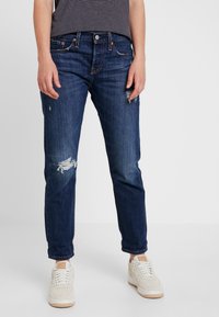 Jeans in denim blu scuro con vestibilità comoda, caratterizzati da toppe strappate sulle ginocchia, cinque tasche e accessori in rame. Indossati con sneakers bianche.