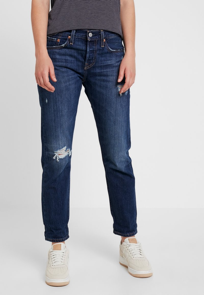 Jeans in denim blu scuro con vestibilità comoda, caratterizzati da toppe strappate sulle ginocchia, cinque tasche e accessori in rame. Indossati con sneakers bianche.