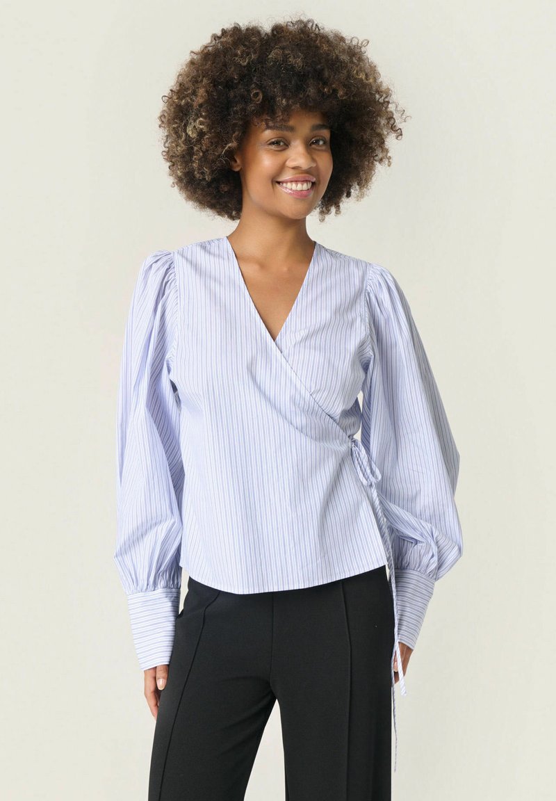 Blusa avvolgente a righe blu chiaro con maniche lunghe a sbuffo e vita annodata, realizzata in tessuto morbido, abbinata a pantaloni neri attillati.