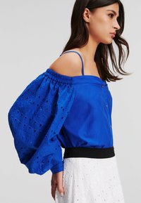 Top blu off-shoulder con maniche a sbuffo testurizzate e motivi a taglio, con una vita nera aderente, impreziosito da bottoni sottili.