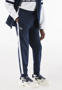 Granatowe spodnie dresowe wykonane z miękkiego materiału, z zielonym logo Lacoste, białymi paskami po bokach oraz elastycznymi mankietami, zestawione z białymi i czarnymi sneakersami.