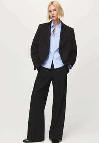 Čierne šité sako cez svetlomodrú košeľu na gombíky, doplnené o čierne široké nohavice a čierne topánky, na neutrálnom pozadí.