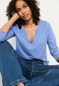 Light blue, long-sleeve shirt with a V-neck and button detail, paired with dark blue wide-leg jeans. Soft texture and casual fit.