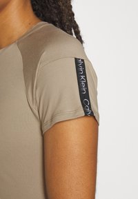 Beżowa koszulka z krótkim rękawem o gładkiej fakturze. Na rękawie znajduje się czarne logo, podkreślające detale marki i szwy.