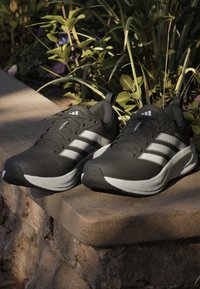 Paire de chaussures de course noires et blanches posées sur un rebord en pierre avec des plantes vertes en arrière-plan.