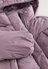 Valge lillaka tooniga polsterdatud jakk, mis on valmistatud pehmest kortsus kangast. Omab tõmbluku- ja trukikinnitust. Paksud, tekstuuriga varrukad.