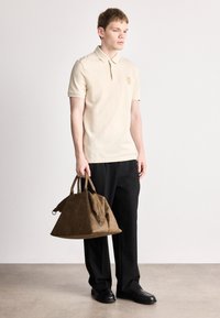 Μπεζ πόλο μπλουζάκι με κοντά μανίκια και γιακά, συνδυασμένο με μαύρο παντελόνι. Κρατά μια καφέ, υφασμένη δερμάτινη τσάντα με λαβές.