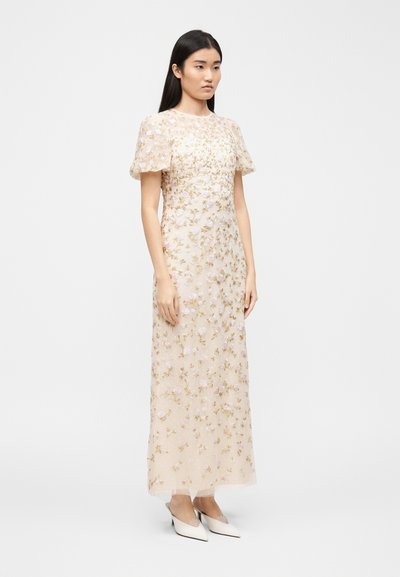 Γυναίκα που φοράει μακρύ φόρεμα εκρού με εμφανείς κοντές φουσκωτές μανσέτες και περίπλοκη κεντητική με λουλούδια, συνδυασμένο με λευκά μυτερά παπούτσια.