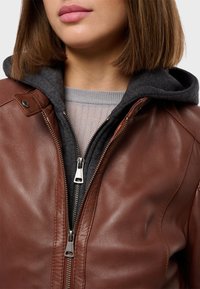 Giacca in pelle marrone con silhouette aderente, caratterizzata da una chiusura con zip argentata, felpa grigia sotto e texture a coste sul collo.