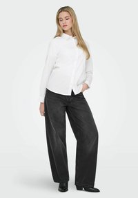 Jeune femme aux longs cheveux blonds portant une chemise blanche boutonnée, un jean noir à jambes larges et des bottes à talons noires, posant avec une main dans la poche.