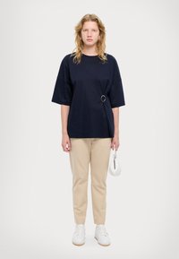 Marineblå oversized t-skjorte med ringdetalj på siden, parret med beige bukser og hvite sneakers, holder en liten hvit håndveske.