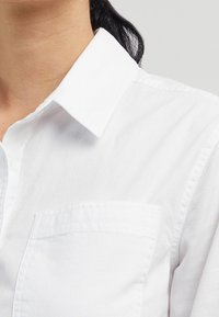 Chemise blanche boutonnée en coton doux avec un col classique et une poche poitrine unique. Caractérisée par une texture lisse et des lignes épurées.