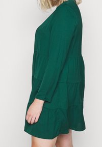 Abito verde a livelli realizzato in un tessuto morbido, con maniche lunghe e una vestibilità comoda. Il design include dettagli arricciati alle cuciture.