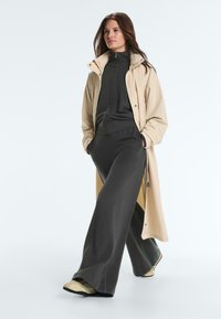 Femeie care poartă o haină lungă bej peste o jachetă neagră cu fermoar și pantaloni largi, purtând adidași bej, pozând pe un fond simplu și deschis la culoare.