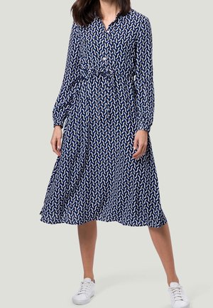 Frau trägt ein blaues, knielanges, langärmeliges Kleid mit weißen geometrischen Mustern und weißen Sneakers, steht mit gespreizten Beinen.