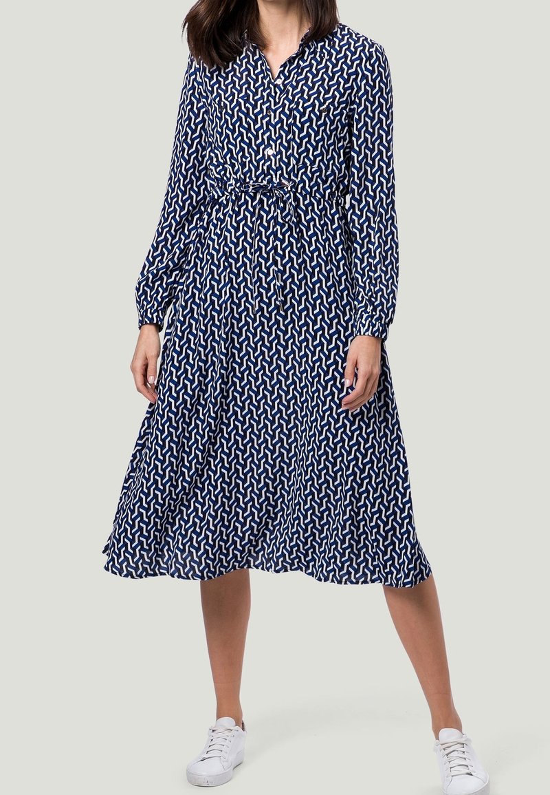 Femme portant une robe bleue à manches longues, longueur genoux, avec des motifs géométriques blancs et des baskets blanches, debout jambes écartées.