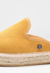 Gul mockaskor med en vävd jutesula, avslutade med en svart logotypdetalj och en kontrasterande sydd kant.