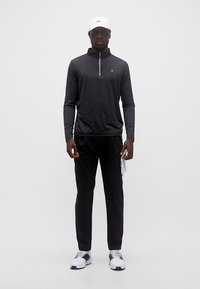 Άνδρας που φοράει μαύρο μακρυμάνικο πουκάμισο με φερμουάρ, μαύρο παντελόνι, λευκό καπέλο, κρατάει γάντι γκολφ και φοράει λευκά αθλητικά παπούτσια, στέκεται μπροστά σε απλό φόντο.