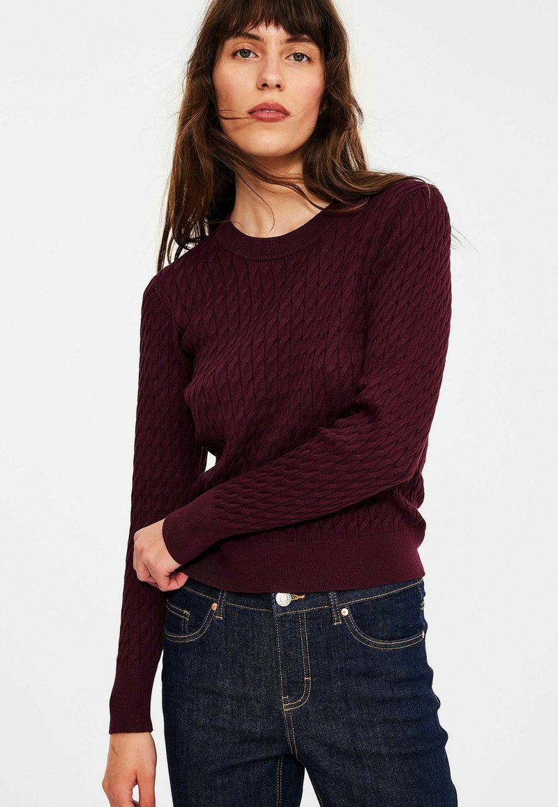 Bordeaux gebreide trui met een gestructureerd ruitpatroon, ronde hals en geribde manchetten. Gecombineerd met donkerblauwe jeans met contrasterende stiksels.