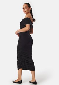Robe noire à décolleté asymétrique avec des détails froncés, longueur genou, silhouette ajustée et texture de tissu lisse. Chaussures plates noires.