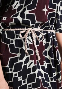 Tmavě vzorované šaty s vínovými a černými abstraktními tvary na světlém pozadí, stažené zaplétaným béžovým opaskem v pase.