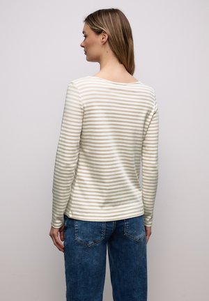 Frau mit glattem hellbraunem Haar, die ein beige-weiß gestreiftes Langarmshirt und blaue Jeans trägt, von hinten vor einem einfarbigen Hintergrund.